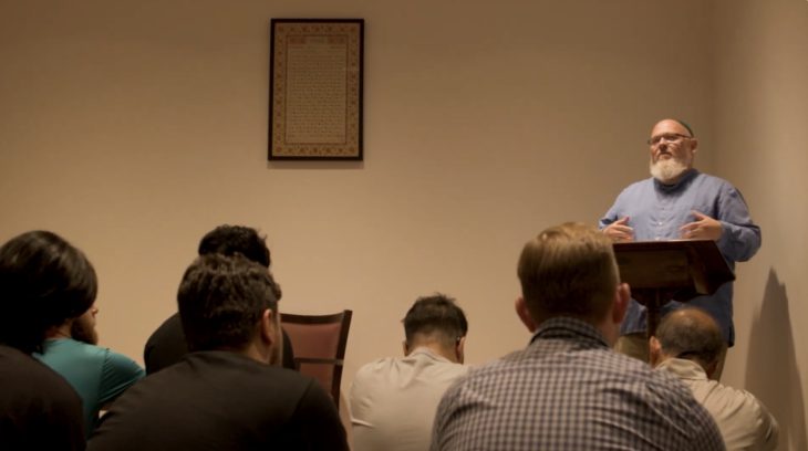 speaker addresses a seated group during the Muslim Life welcome dinner, standing at a wooden podium in a quiet room with framed art on the wall.